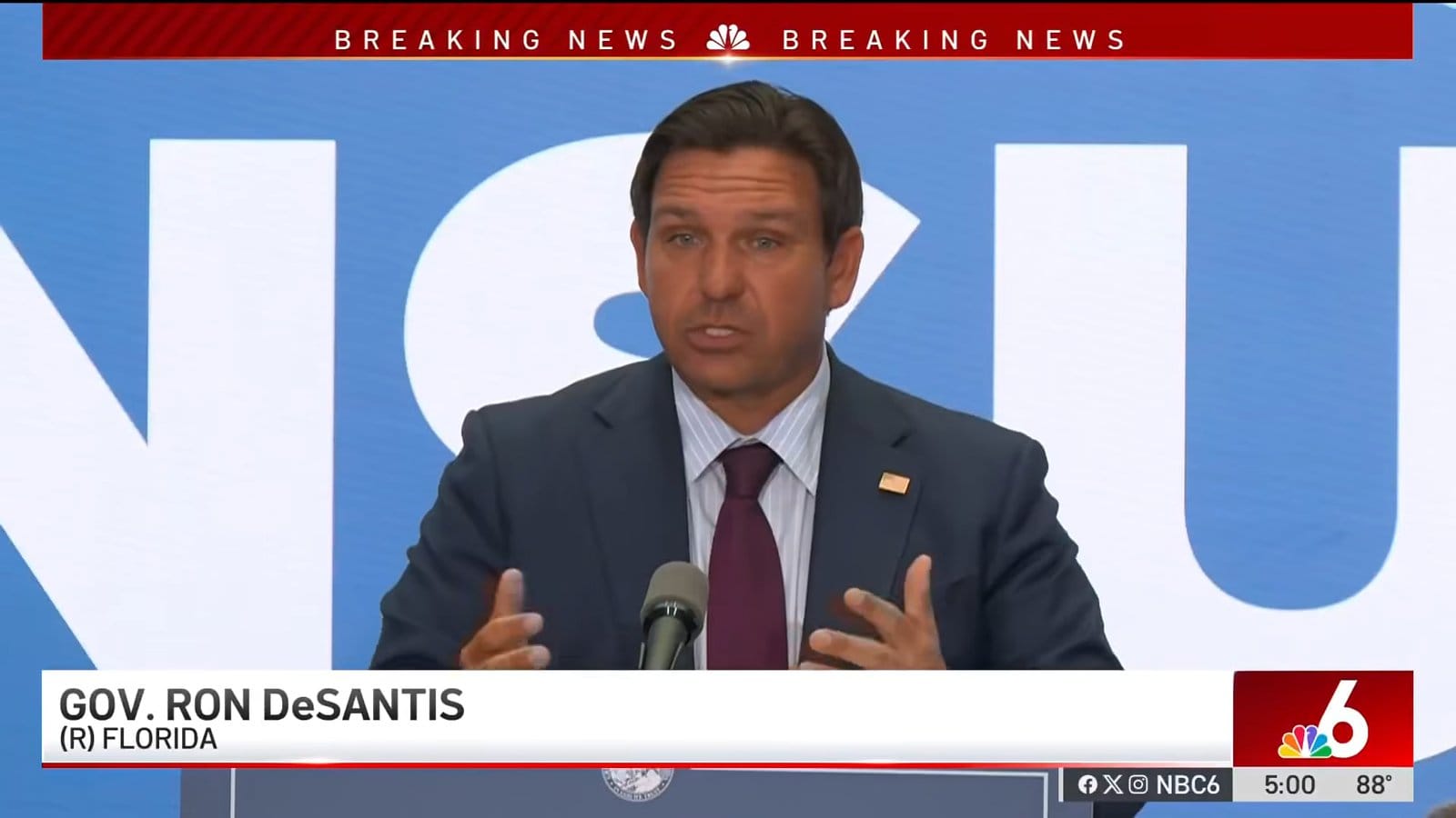 Governador Ron DeSantis fala ao microfone em coletiva, com tarja da NBC6 identificando seu nome e cargo
