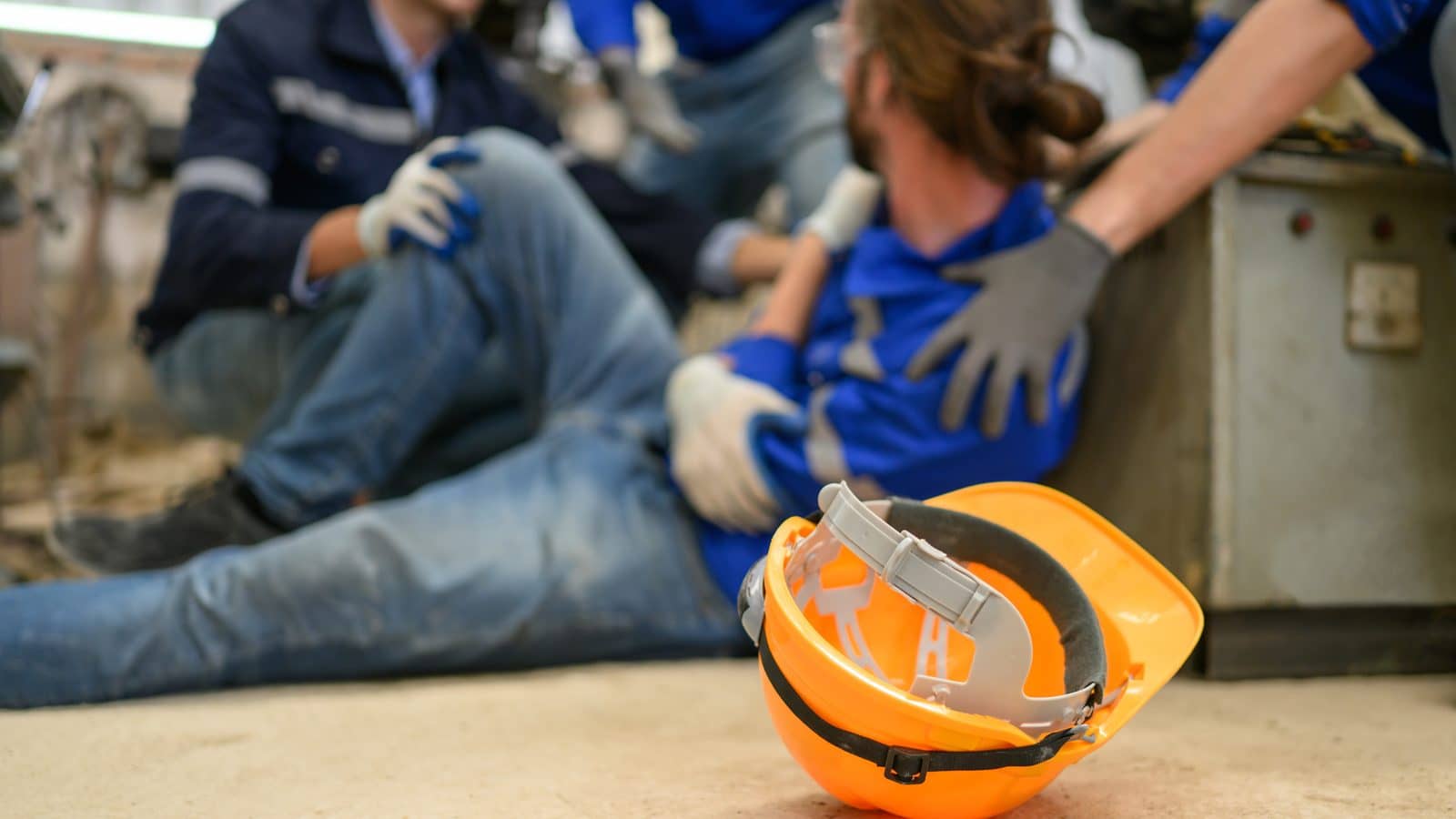 Trabalhador acidentado sentado no chão sendo amparado por colegas, com capacete laranja em primeiro plano