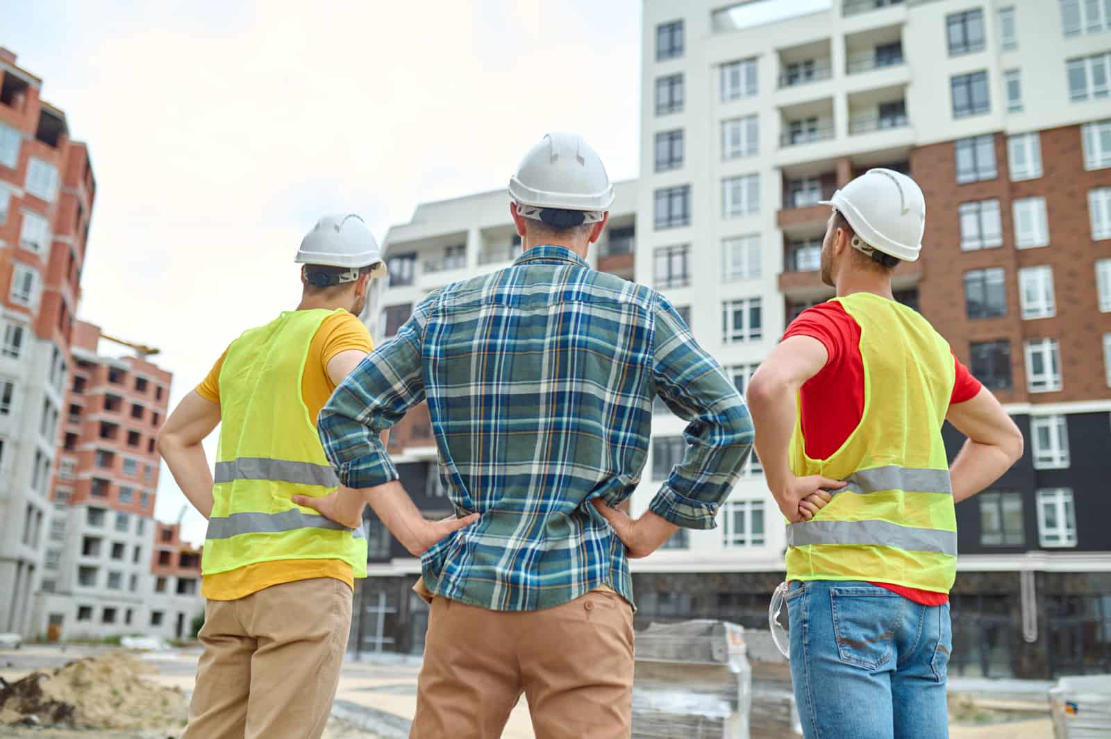 Três trabalhadores da construção civil de capacete observando prédio em construção