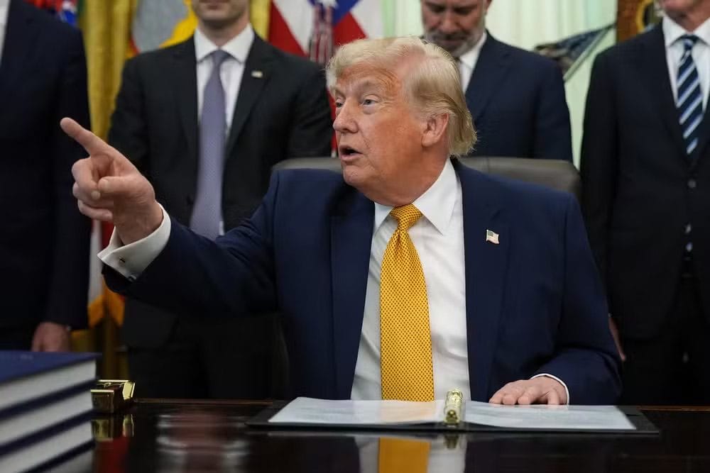 Donald Trump sentado à mesa no Salão Oval, gesticulando durante assinatura de documentos, cercado por assessores
