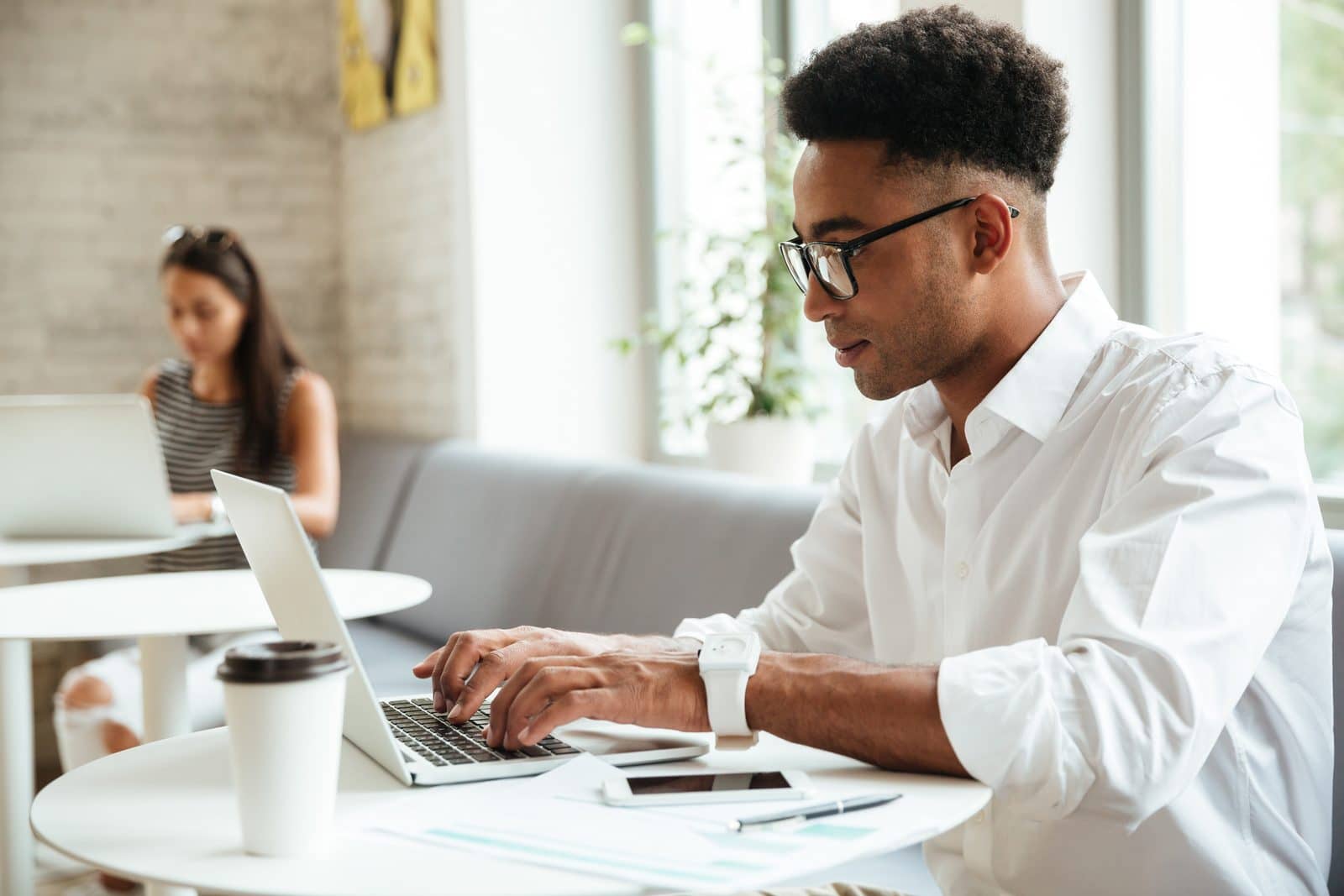 Homem de óculos e camisa branca digita em laptop em espaço de coworking, com outra pessoa trabalhando ao fundo