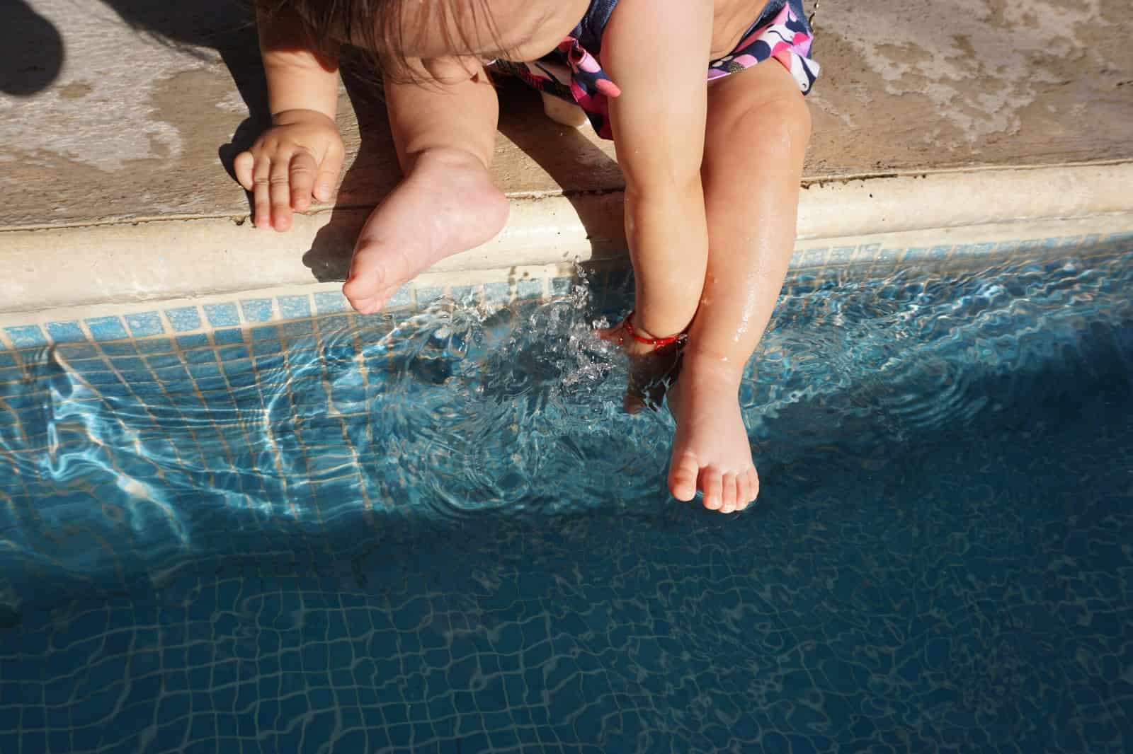 Criança pequena sentada na borda de uma piscina com os pés na água, sem adulto ao alcance