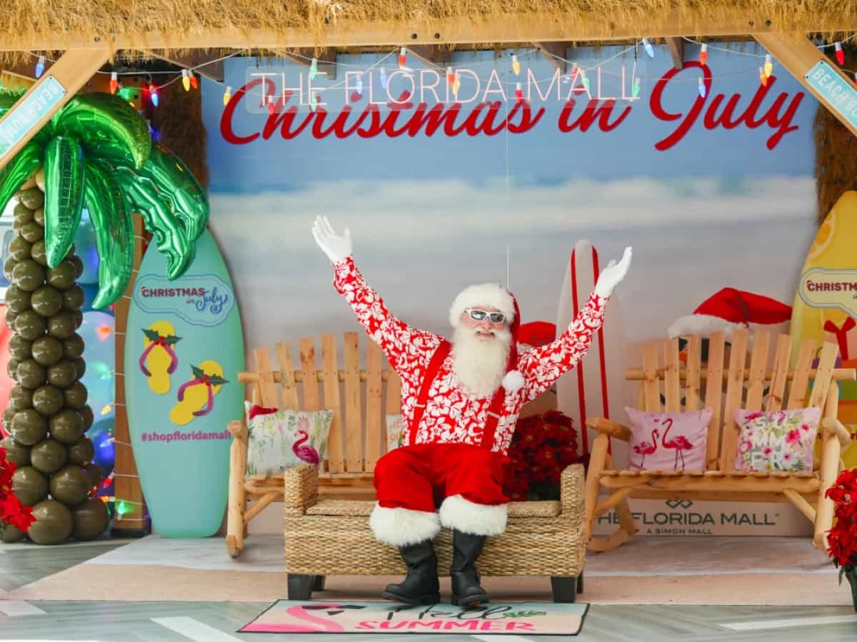 Papai Noel de camisa havaiana vermelha e óculos escuros abre os braços em cenário praiano do Christmas in July no Florida Mall