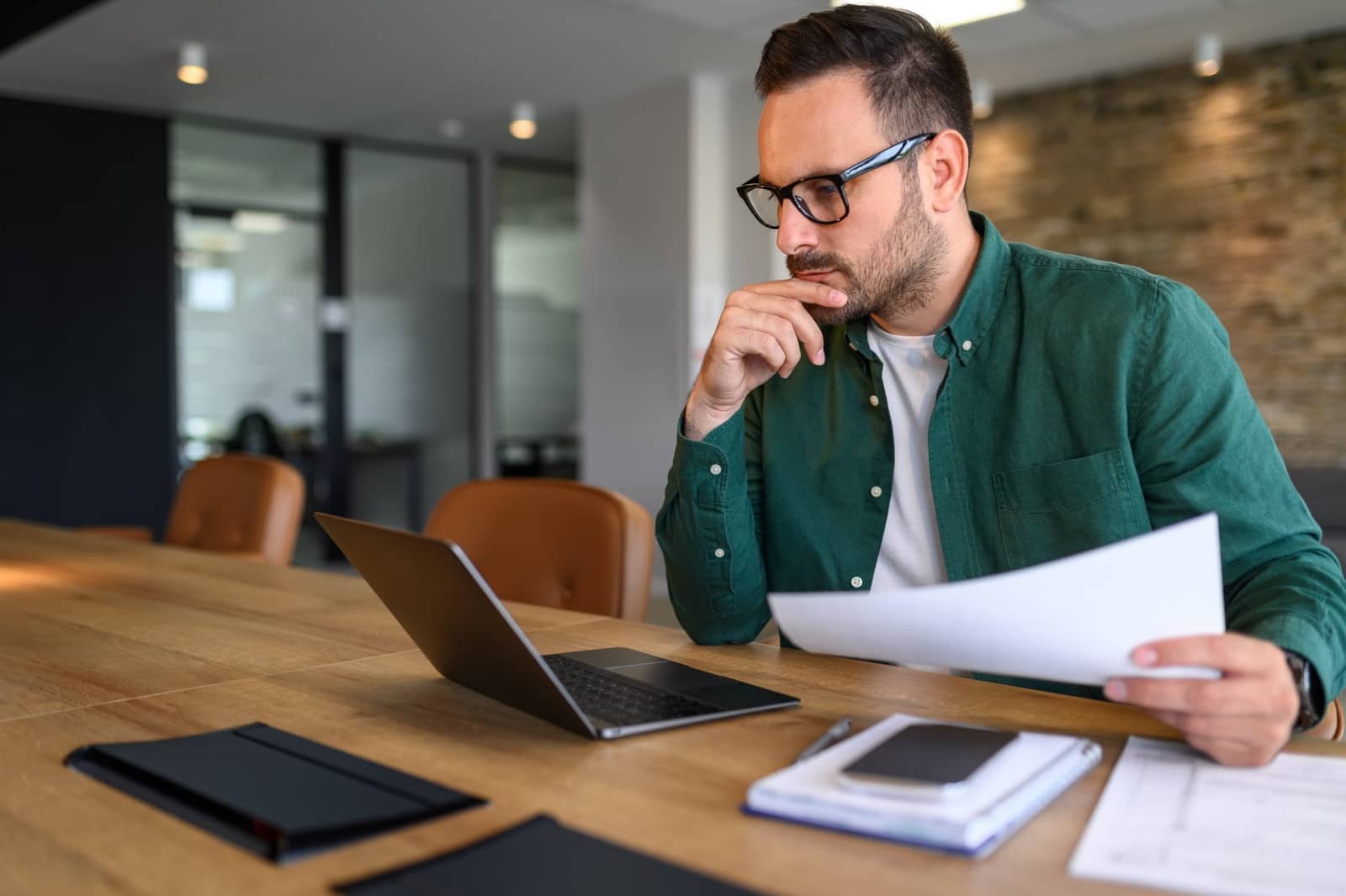 Homem de óculos analisando documentos ao lado de um laptop em sala de reunião