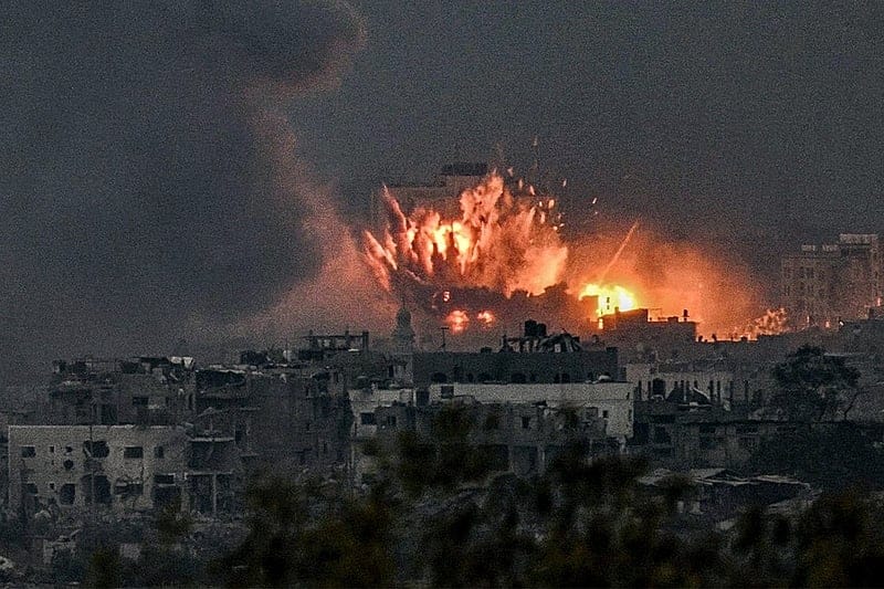 Cidade sob bombardeio ao entardecer, com explosões e labaredas de fogo entre prédios destruídos e uma coluna de fumaça escura no céu