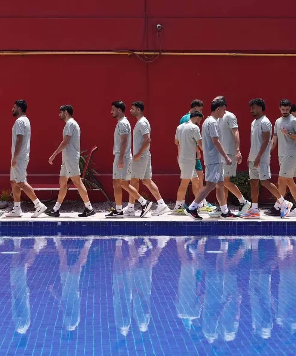 Jogadores da seleção do Irã caminham em fila ao redor da piscina de um hotel, diante de uma parede vermelha