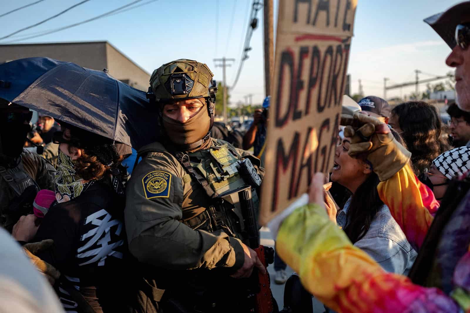 Agente federal em traje tático cerca manifestantes, um deles com cartaz contra deportações