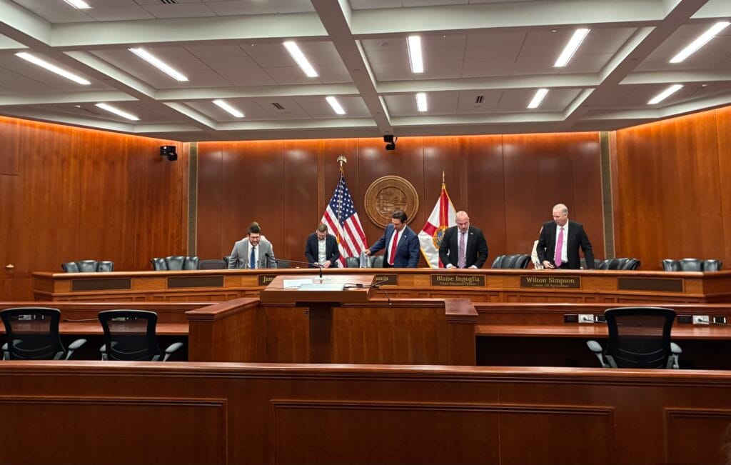Governador Ron DeSantis e membros do Gabinete da Flórida em pé diante das bandeiras dos EUA e do estado, em sala de reunião