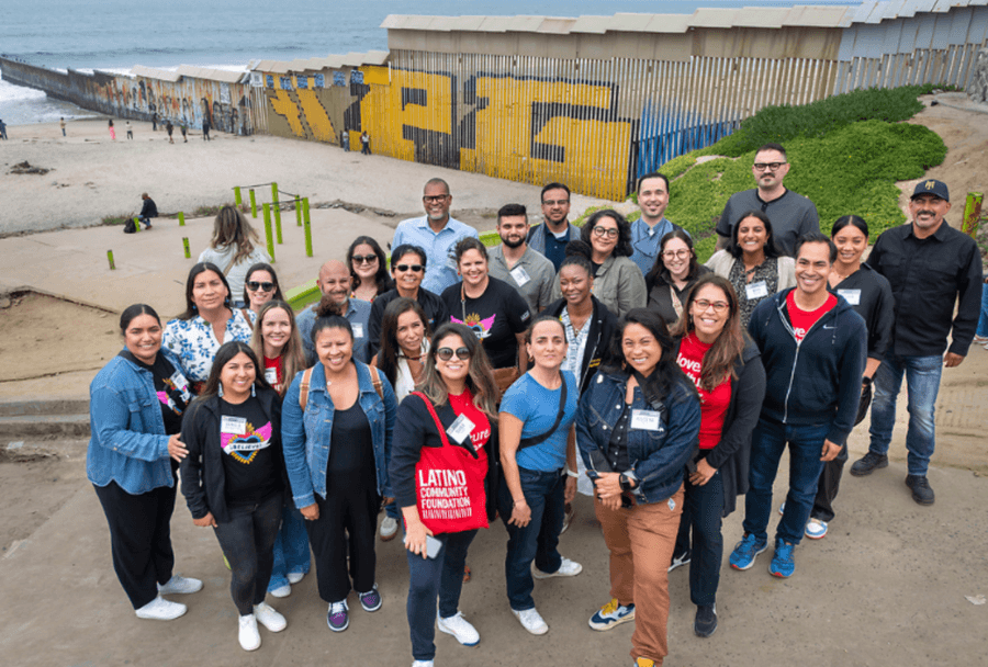 Grupo de participantes da Latino Community Foundation posa diante do muro da fronteira EUA-México, um deles segurando bolsa com o nome da fundação