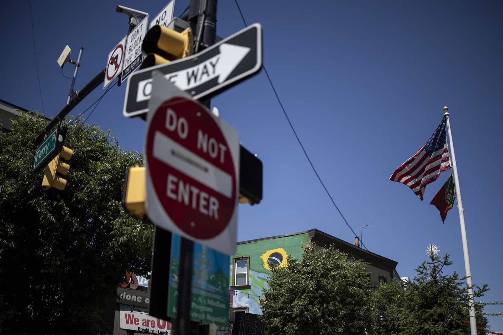 Cruzamento de rua com placas de trânsito, bandeira dos EUA e mural em verde e amarelo ao fundo, em bairro de imigrantes