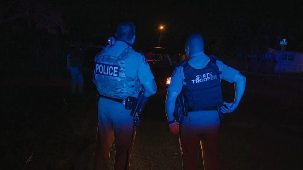 Dois agentes vistos de costas à noite, um com colete escrito POLICE e outro STATE TROOPER, em via mal iluminada