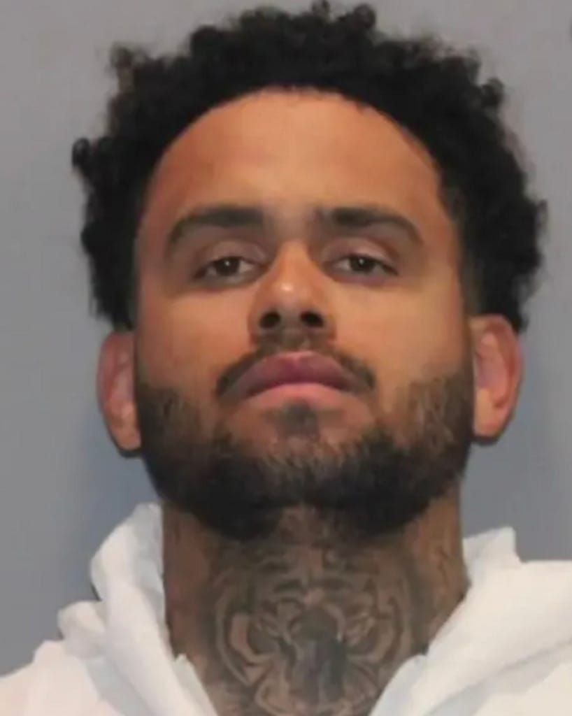 Foto de identificação policial de um homem jovem, de barba e cabelo curto encaracolado, com tatuagem no pescoço