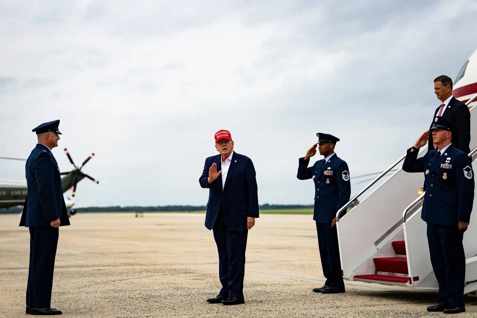 O presidente Donald Trump, de boné vermelho, ergue a mão diante de um oficial da Força Aérea enquanto dois militares o saúdam junto à escada de um avião