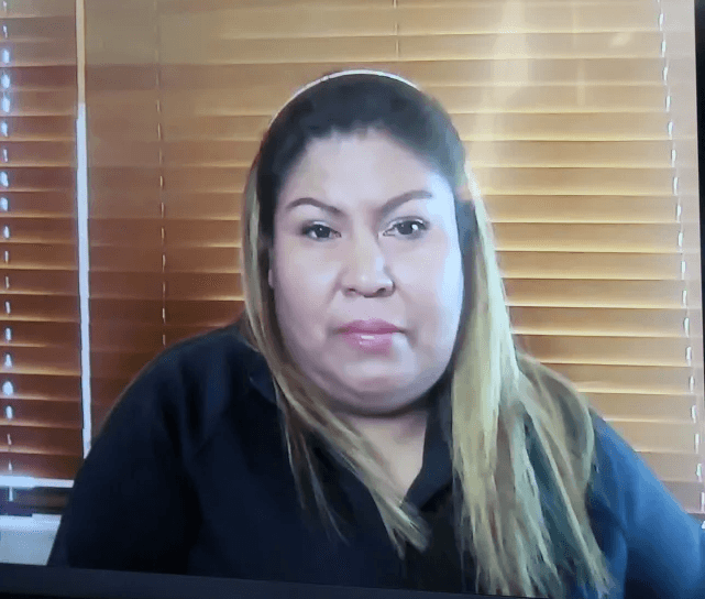 Mulher de cabelos castanhos, vista de frente diante de uma janela com persianas, durante entrevista