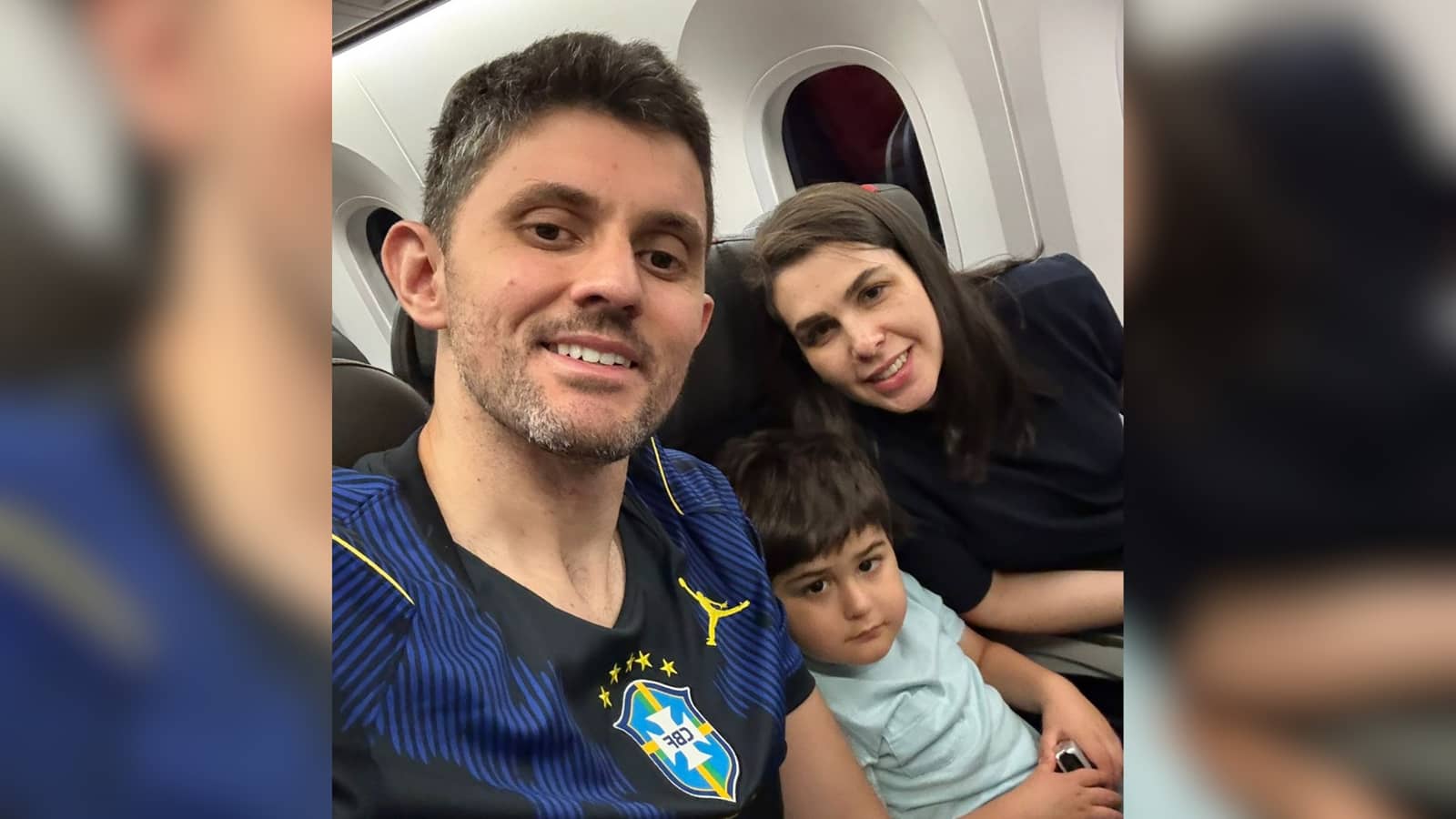 Casal e uma criança pequena fazem selfie sentados em um avião; o homem veste camisa azul da seleção brasileira