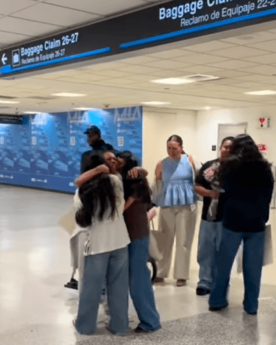 Família se abraça em um saguão de desembarque de aeroporto, próximo a placas de retirada de bagagem em inglês e espanhol