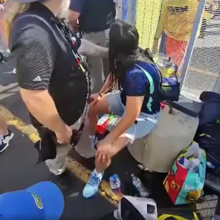 Mulher sentada em uma mureta perto do gradil de um estádio é amparada por um homem que se agacha ao seu lado, cercada por sacolas e garrafas de água