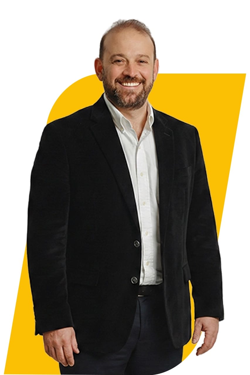Retrato do empresário Alex Colombini, homem de barba grisalha, blazer escuro e camisa branca, sorrindo diante de fundo branco com um grafismo amarelo