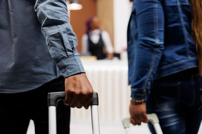 Duas pessoas puxam malas de rodinha em um saguão, vistas da cintura para baixo, com um balcão de recepção desfocado ao fundo