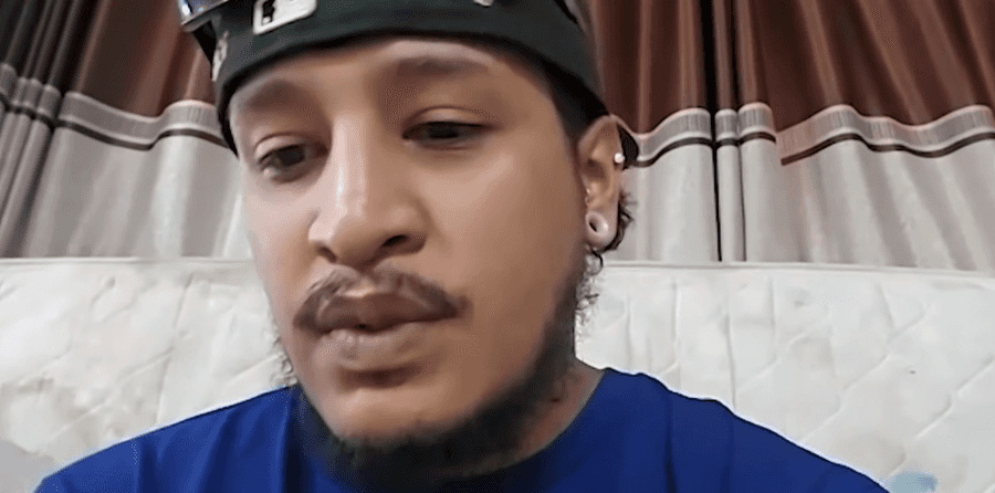 Homem jovem de boné e camiseta azul fala olhando para a câmera, em um cômodo com cortinas claras ao fundo