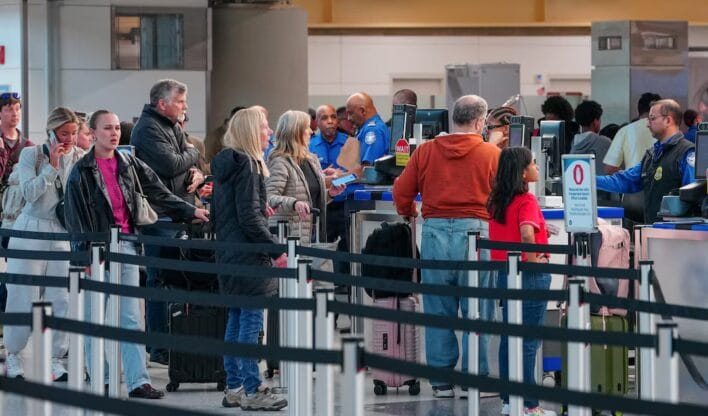Fila de passageiros de diferentes idades aguarda na inspeção de segurança de um aeroporto, com agentes da TSA ao fundo