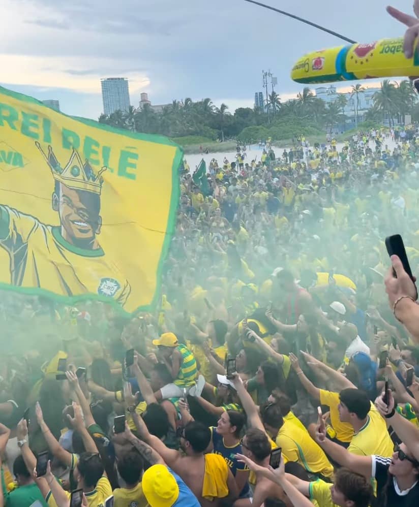 Multidão de torcedores vestidos de verde e amarelo lota a areia de Miami Beach, com uma grande bandeira do 'Rei Pelé' e fumaça verde, sob palmeiras e prédios ao fundo