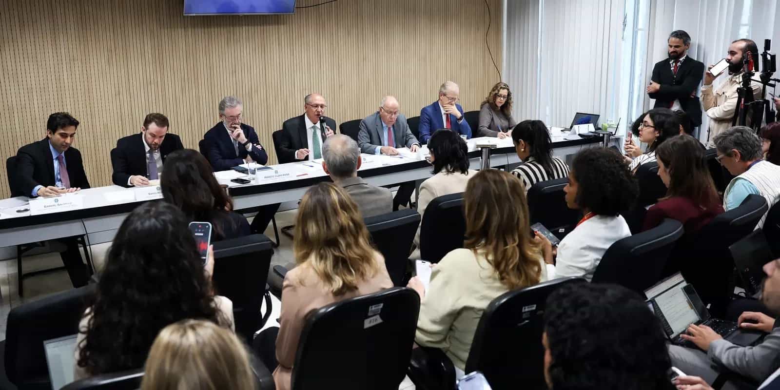 Autoridades do governo brasileiro sentadas a uma mesa durante reunião com o setor produtivo, diante de jornalistas com celulares e laptops