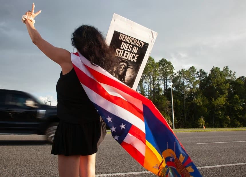 Manifestante de costas, com uma bandeira dos Estados Unidos sobre os ombros, faz sinal de paz à beira de uma rodovia enquanto um veículo passa; ao fundo, um cartaz com os dizeres 'Democracy dies in silence'