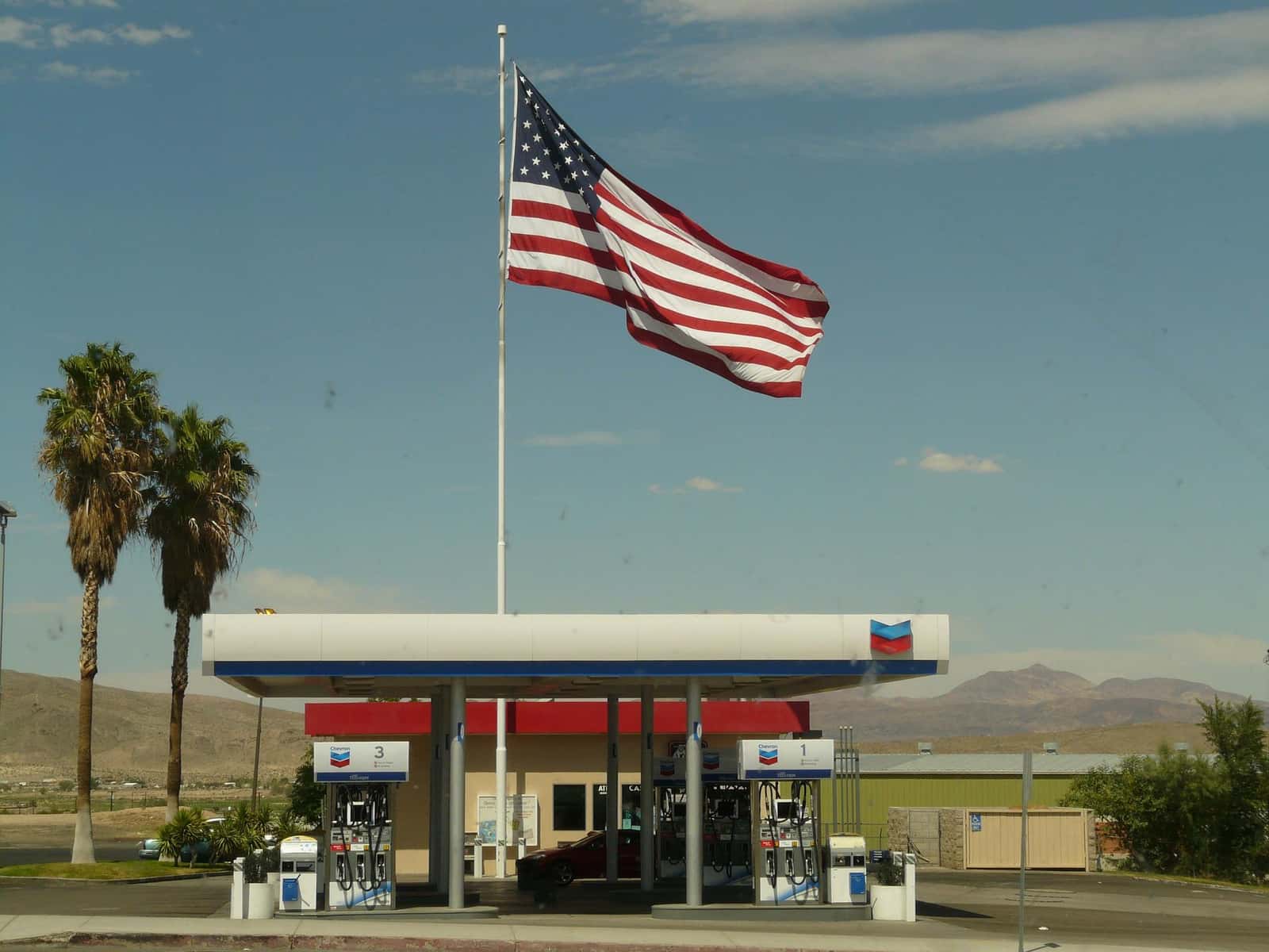 Posto de combustível com grande bandeira dos Estados Unidos hasteada, palmeiras à esquerda e montanhas desérticas ao fundo