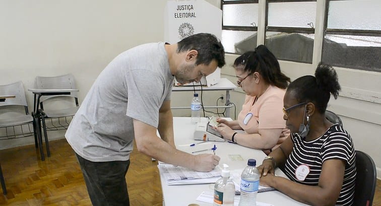 Eleitor assina a lista de presença diante de uma urna e de duas mesárias em uma seção eleitoral, sob o cartaz da Justiça Eleitoral