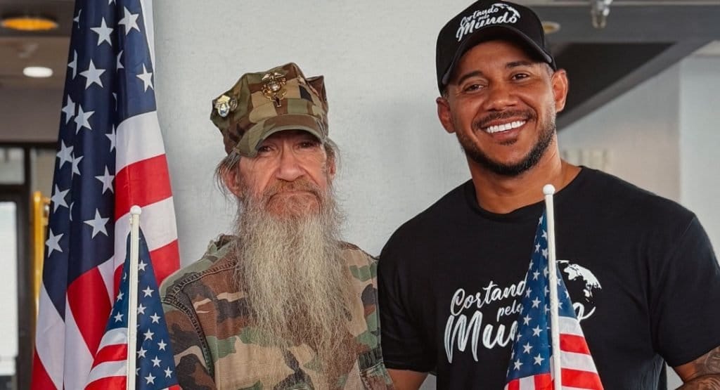 Barbeiro brasileiro Diego Henrique, com camiseta e boné do projeto Cortando pelo Mundo, ao lado de veterano americano fardado entre bandeiras dos EUA
