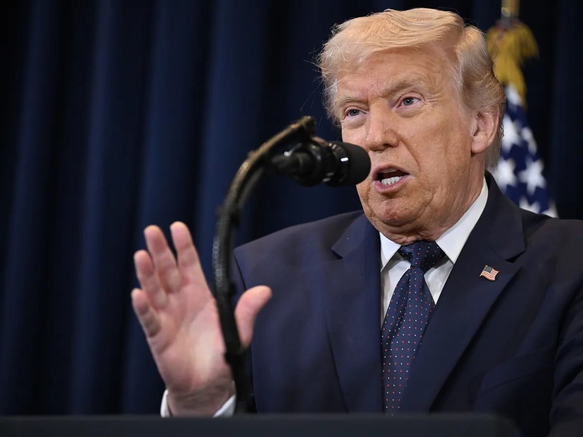 Donald Trump fala ao microfone diante de cortina azul e bandeira dos EUA